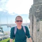 Alex Theophilus sat on a wall wearing a green jumper, sunglasses and a backpack. In the background there is water and boats.