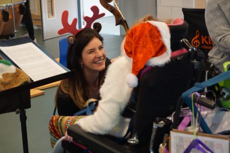 Female violinist with long brown hair in reindeer antlers kneeling in front of a child in a wheelchair wearing a santa hat. The violinist is showing the child her violin.