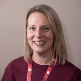 Abi is wearing a maroon Music Mark t-shirt and lanyard. She has blonde hair and is smiling at the camera.