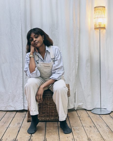 Marie Bashiru, a recipient of the Alan Surtees Trust in 2021, sits on a stool looking at the camera in dungarees and socks