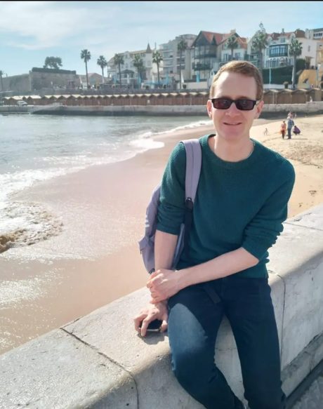 Alex Theophilus sitting on a wall overlooking a beach and water. He has short blonde hair, is wearing sunglasses, a green jumper and a backpack on one shoulder