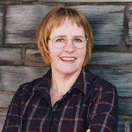 Portrait photo of Asa in glasses, looking towards camera smiling. She is standing in front of a grey brick wall.