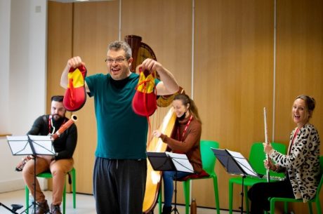 A man is stood holding two large clown shoes. A bassoon player, harpist, and flautist are sat in the background smiling.