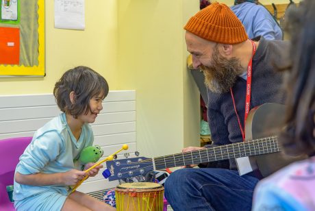 A man with a beard and orange hat is holding a guitar towards a child, who has a percussion instrument in their hand