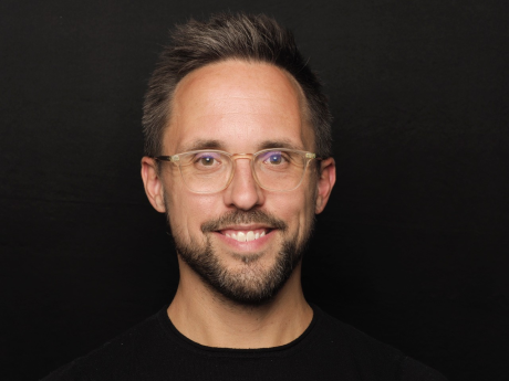 Dr Axel Petri-Preis smiling, wearing a black t-shirt in front of a black background. He has short brown hair, facial hair and glasses.