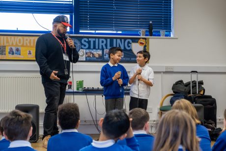 Two school children stood holding microphones with a rapper in front of a group of children sat watching.
