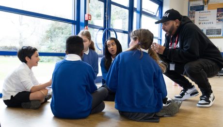 Five school children are sat on the floor talking, a rapper is crouching down speaking to them.