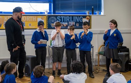 Five children stood holding microphones rapping in front of other children sat watching. A man is also stood at the front leading the session.