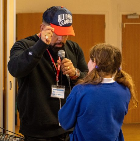 A rapper wearing a black sweatshirt and baseball cap reading 'Billionaire boys club'. He is holding a microphone and is facing a young student with her back to the camera.