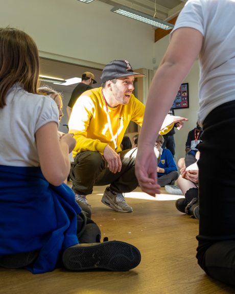 A rapper crouched down working with a group of children. He is wearing a yellow sweatshirt and a black baseball cap, dark trousers and trainers. Children can be seen around him.