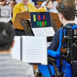 Clarion player in rehearsal. He is a wheelchair user, and has a windows device on his music stand, with his music sheet on the keyboard. He is wearing a Quha Zono headset, with which he controls the notes on his computer screen, which are represented in colour blocks.