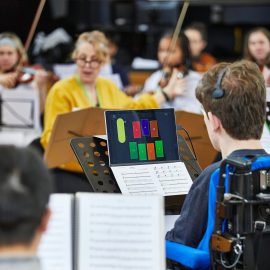 Clarion player in rehearsal. He is a wheelchair user, and has a windows device on his music stand, with his music sheet on the keyboard. He is wearing a Quha Zono headset, with which he controls the notes on his computer screen, which are represented in colour blocks.