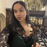 Selfie of Danya with long black hair, wearing a black top and holding a viola