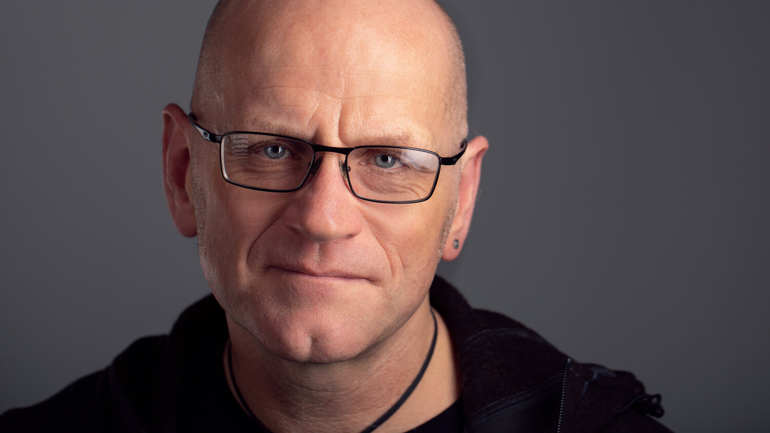 Dave is wearing glasses, he is wearing a black top. He has a bald head and a black necklace.