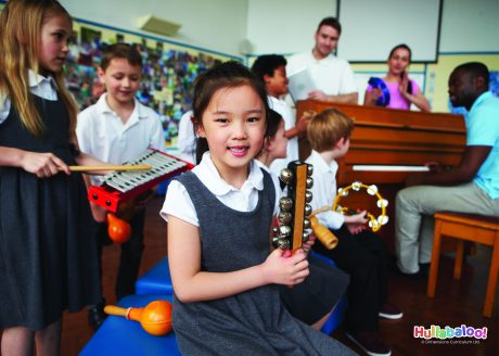 Child holding bells smiling at the camera, with more children in the background holding percussion instruments and teachers stood round a piano