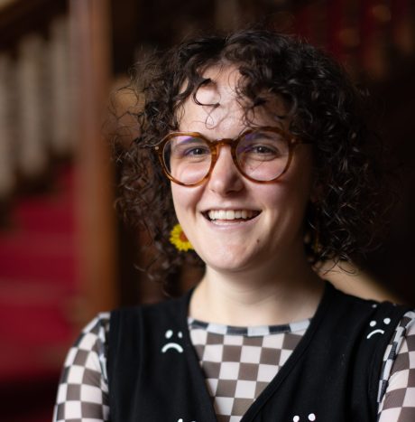 Hannah is wearing a checkered long-sleeve top with a black vest over the top with smily faces on it. They have short curly hair, are wearing glasses and smiling.