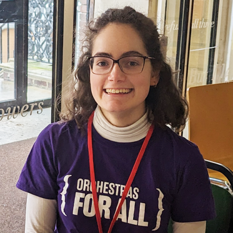 Helen has brown hair and glasses and is smiling at the camera. She is wearing a purple t-shirt reading 'Orchestras for all'.