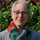 Helen has short grey hair and glasses. She is wearing a colourful scarf and is stood outside smiling.