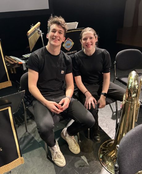 Holly is wearing all black and is sat next to another player from Grimethorpe Colliery Band. Chairs and some instruments can be seen around them. They are both smiling at the camera.