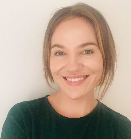 a woman with dark blonde hair and a dark green top is smiling at the camera, against a white wall