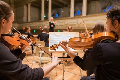 Two violinists playing with a music stand in front of them and a conductor at the front, taken from behind the violinists' heads.