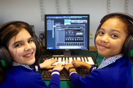 Two children wearing headphones smiling at the camera. They are using a MIDI keyboard plugged into a computer with music tech software on the screen.