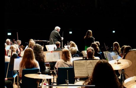 The back of an orchestra with several music stands and a conductor and soloist stood at the front