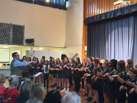 A group of school children stood singing and doing hand gestures with a conductor facing them, and people sat in the audience