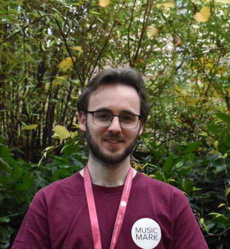 Matthew Crisp wearing a Music Mark t-shirt and glasses smiling at the camera, with foliage in the background.
