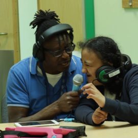 Teacher with headphones on holding microphone to young persons mouth. They are also wearing headphones.