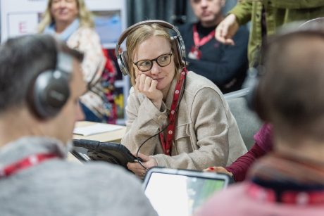 A woman sat holding a tablet and wearing headphones and glasses, resting her chin on her hand, looking to the side. Other people can be seen around the side of her, blurry, some also wearing headphones.