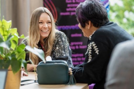 Two women sat at a table talking, one woman is only visible from the back, the other women is smiling.