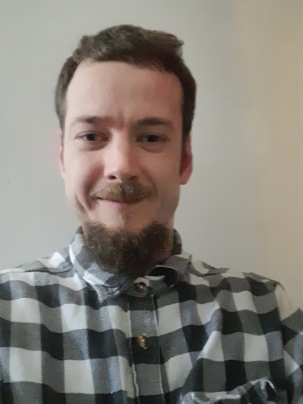 Nathaniel Ford-Francis smiling at the camera. He has short brown hair, a moustache and beard, and is wearing a black and white plaid shirt.