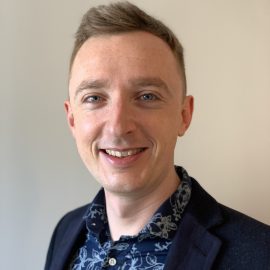 Nick has short hair and is wearing a blue and white patterned shirt and a dark blazer. He is smiling at the camera.