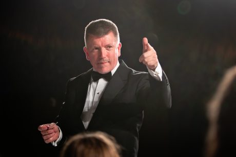 Peter Smalley wearing a suit and bow tie holding a conductors baton and pointing ahead
