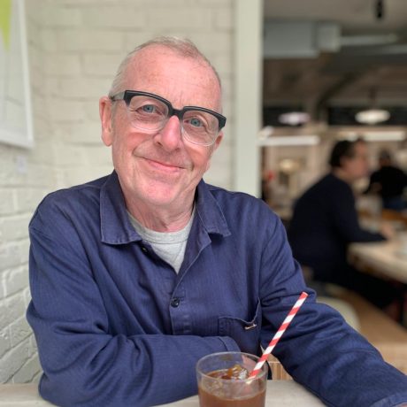 Philip Flood smiling at the camera, wearing a blue shirt over a grey t-shirt and black glasses. He has short hair and a drink on the table in front of him.