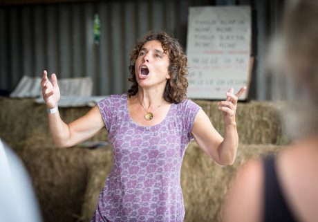 Pili is wearing a purple top and is stood with her arms raised, singing.