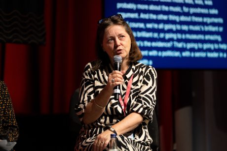 Bridget Whyte say holding a microphone mid-speaking. She is wearing a black and white dress, a red lanyard and has brown, shoulder-length hair