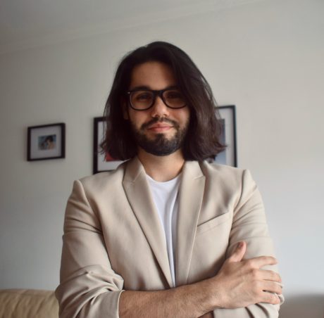 Javier Rivas looking at the camera. He has his arms folded and is wearing a cream blazer with white top. He has long dark hair and glasses.