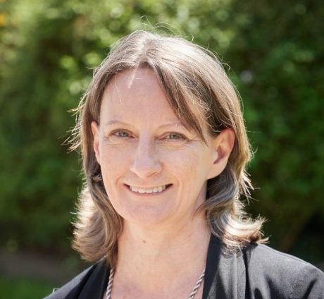 Ruth has shoulder length brown hair and is wearing a black blazer and a patterend top. She is stood outside, smiling at the camera.
