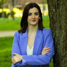 Kitty Bazalgette in blue jacket, leaning on tree with park in the background