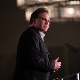 Image of Scott Welcomme talking with a microphone in front of him. He is in a suit and glasses.