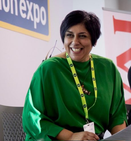 Sharon is wearing a green top, a yellow lanyard and has short black hair. She is smiling past the camera.