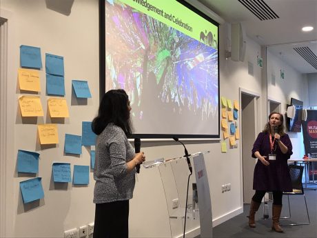 Dr Ayesha Nathoo and Susannah Tresilian present in front of a screen. There are sticky post it notes on the wall behind them 