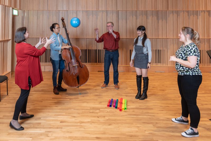 Five musicians are playing in a music education class, someone is throwing a ball, someone is playing a cello, there are boomwhackers on the floor. Two other people wait to catch the ball.
