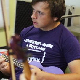 Young Person playing the Ukulele, with young person in foreground playing a stringed instrument. Only the bow and instrument neck are visiable on the violin or cello.