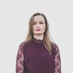 Aileen Sweeney stood with a white background. She is wearing a dark purple top with lace and has long blonde hair