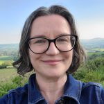 A selfie of Clair with short dark hair and black glasses in front of hilly countryside