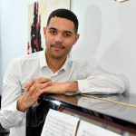 James B. Wilson stood leaning on a piano wearing a white shirt. He has short black hair and some facial hair