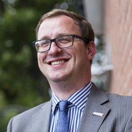 Man in glasses and grey suit smiles towards camera.